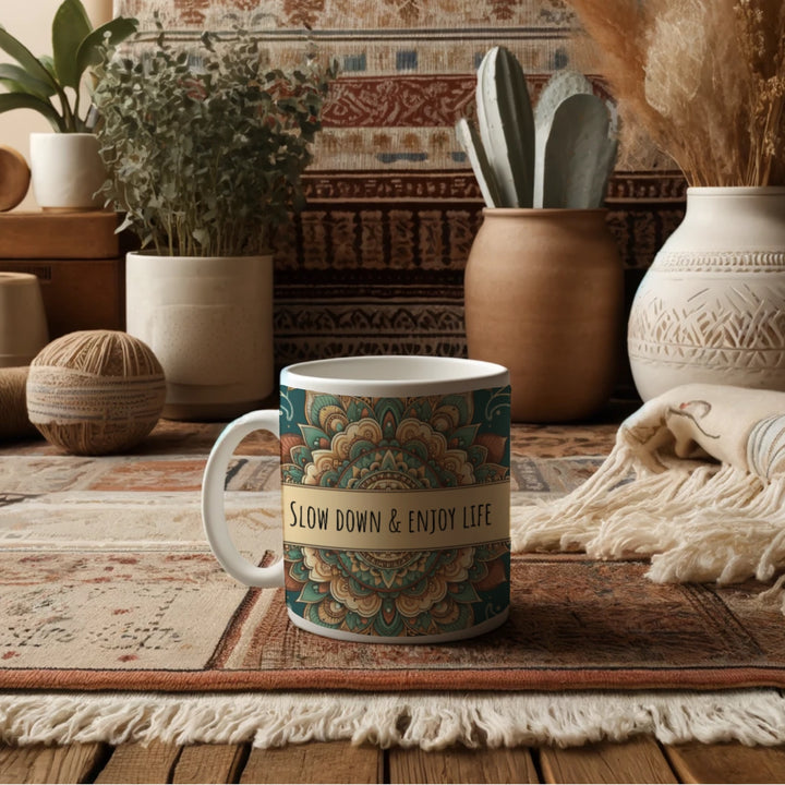 White ceramic mug with a detailed green and beige mandala design and the phrase 'Slow Down & Enjoy Life' in the center, placed in a cozy bohemian-style room with earthy-toned decor, including woven baskets, pottery, patterned rugs, and indoor plants.