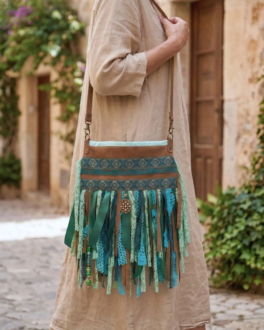Woman wearing a handmade upcycled bohemian crossbody bag in turquoise and green with fringe and lace details, styled with a beige linen dress in a Mediterranean village setting