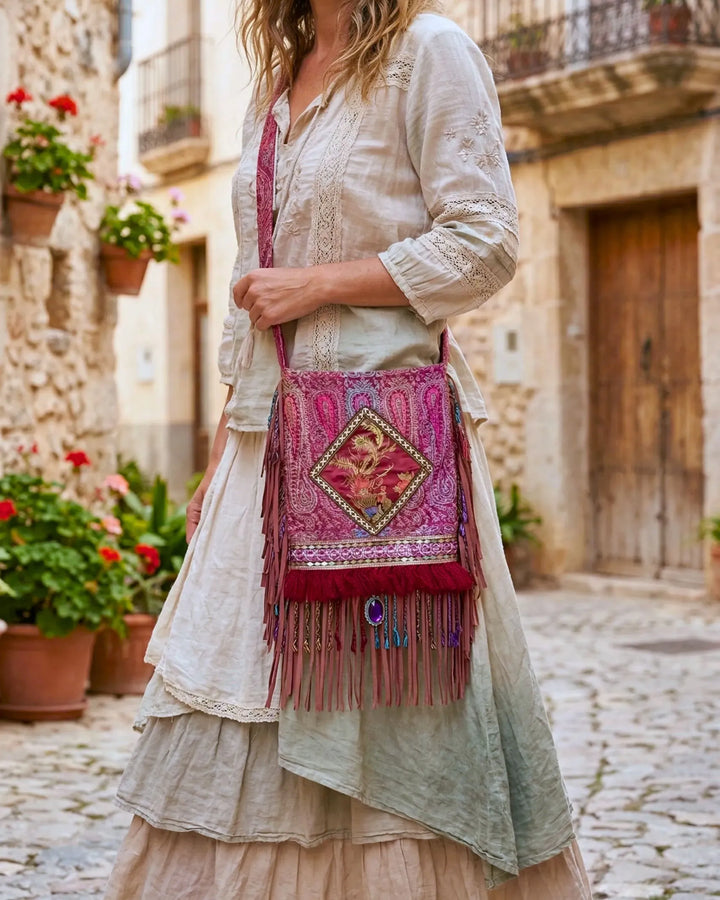 Bohemian upcycled crossbody bag in pink and burgundy tones with embroidered details and long leather fringes, worn by a woman in a flowy linen dress in a Mediterranean street with flowers and stone houses