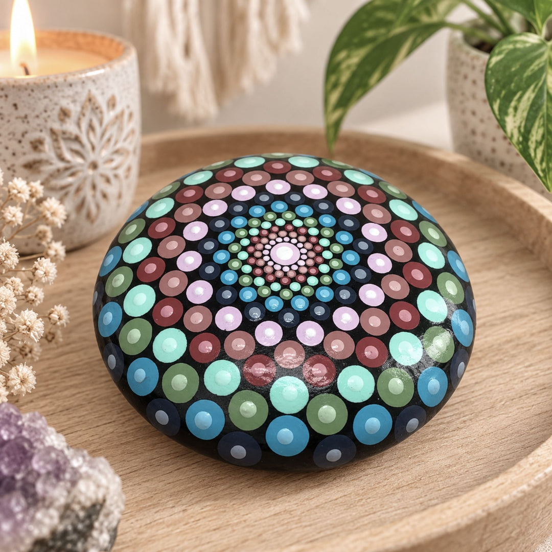 Hand painted mandala stone with colorful dot art in turquoise, blue, pink and green, styled on a wooden tray with candle, crystal and plant.