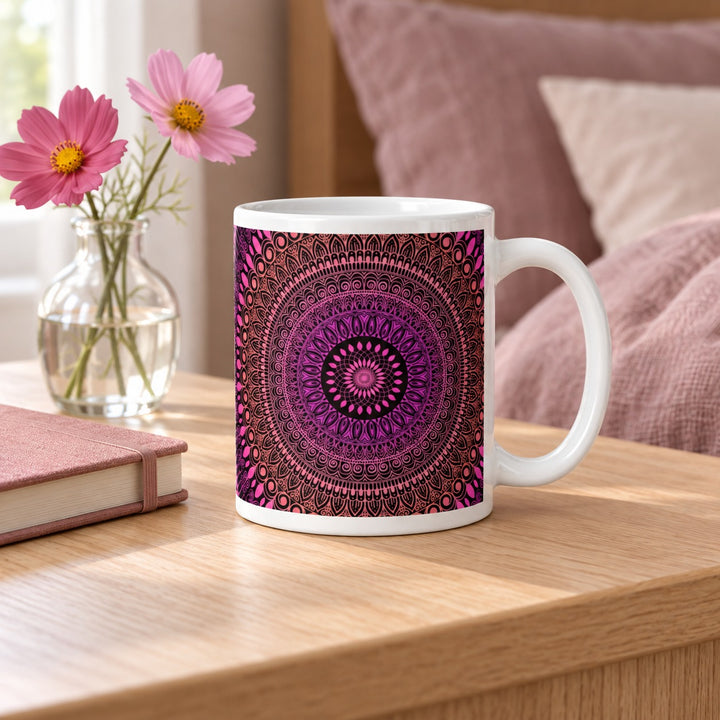 11 oz white ceramic mug with pink, purple and salmon mehndi mandala on a bedside table beside pink flowers.