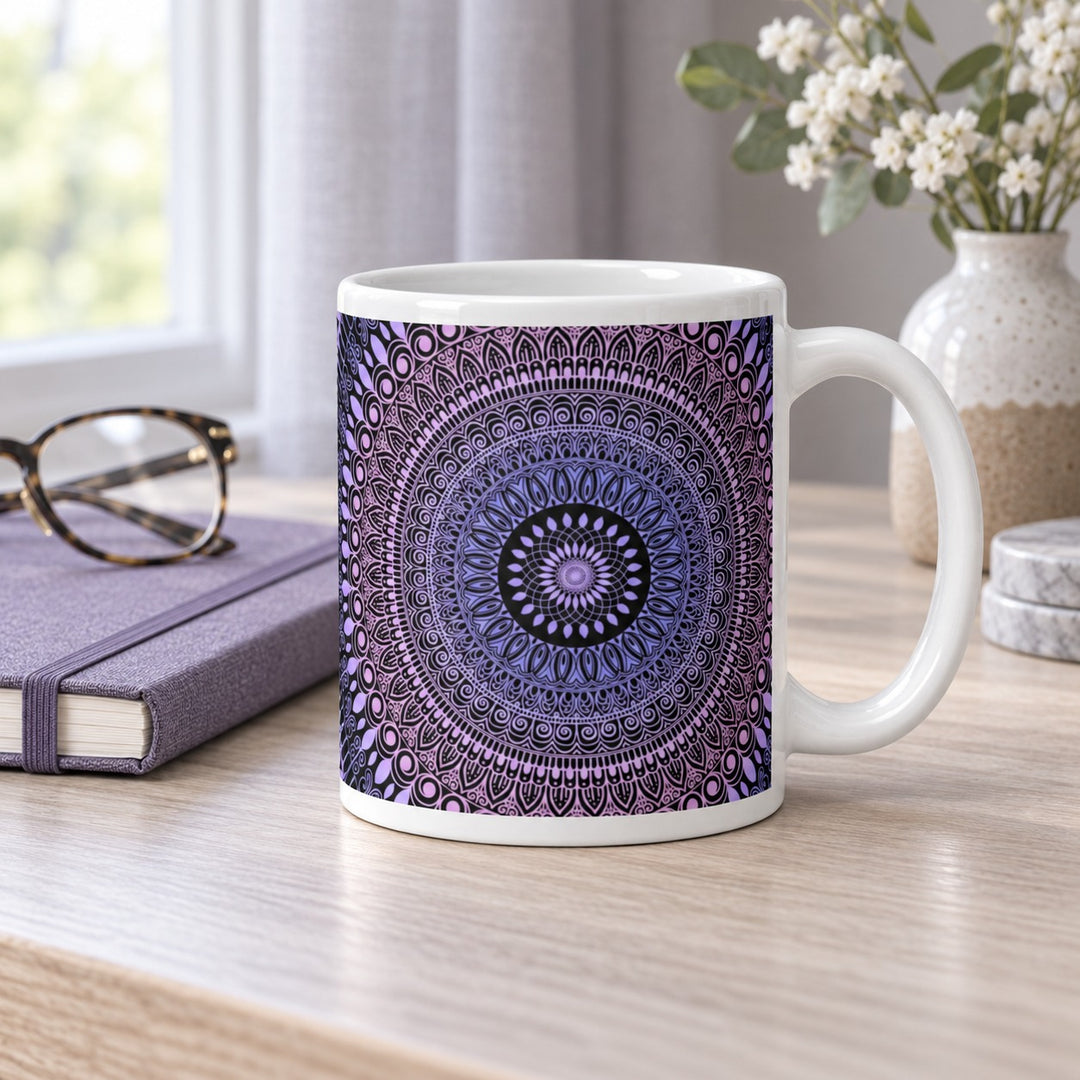 11 oz white ceramic mug with pink and lavender mehndi mandala on a pale wood desk beside a lavender notebook.