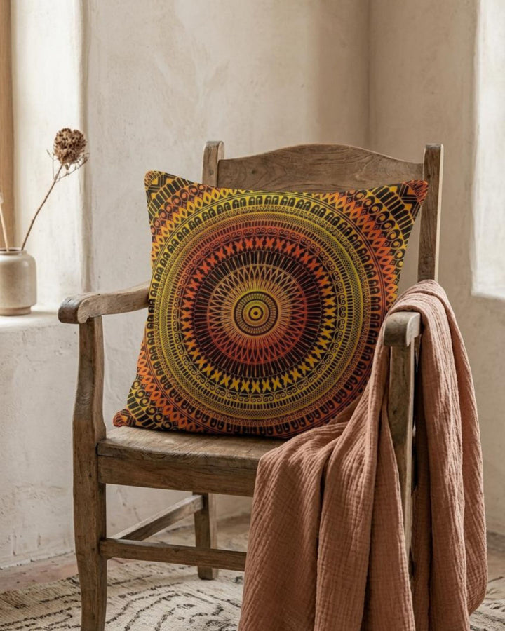 Boho mandala pillow cover with warm orange and black pattern placed on a rustic wooden chair in a soft neutral interior, styled with a textured throw blanket and natural decor in warm earthy tones