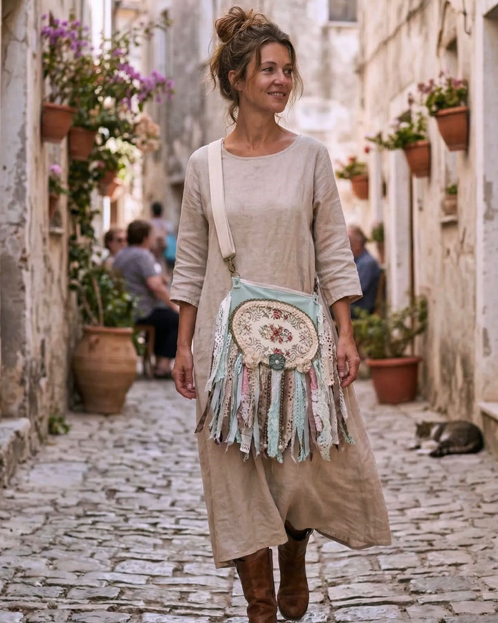 Woman walking through a Mediterranean street wearing a handmade boho crossbody bag with pastel tones, lace and fringe, styled with a beige linen dress and boots